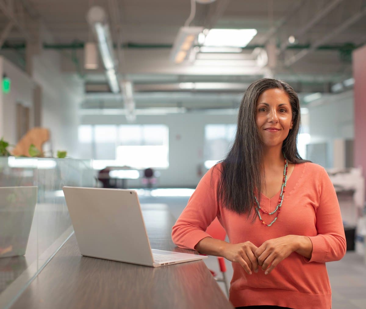 Woman in an orange shirt sitting beside a table with a laptop, working extra hours