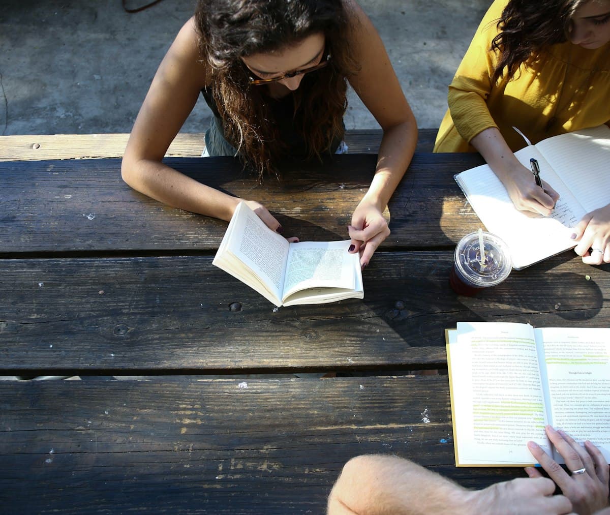Two students reading books and writing in a notebook at a wooden picnic table